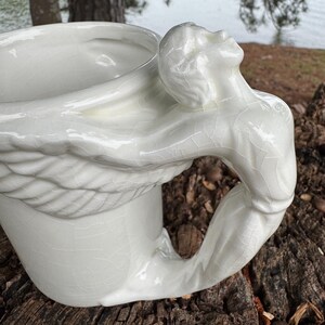 May include: A white ceramic mug with a unique handle sculpted in the form of a human figure. The figure's arm forms the handle, and the head is tilted back. The mug has a crackle glaze finish, adding texture to the design.