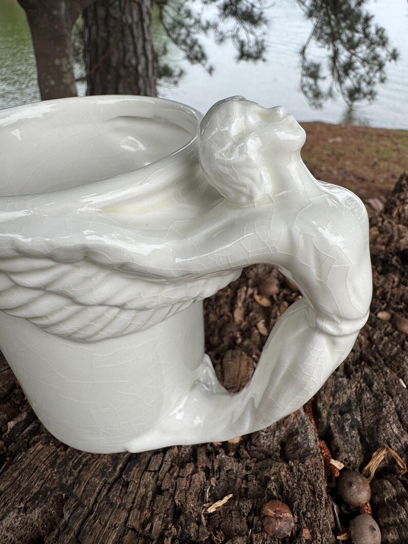 May include: A white ceramic mug with a unique handle sculpted in the form of a human figure. The figure's arm forms the handle, and the head is tilted back. The mug has a crackle glaze finish, adding texture to the design.