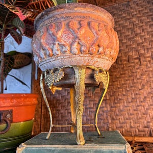 May include: A weathered terracotta planter with a raised scroll pattern sits inside a gold-toned metal stand featuring grape leaf accents. The stand rests atop a green hardcover book titled Principles of College Physics with visible wear.