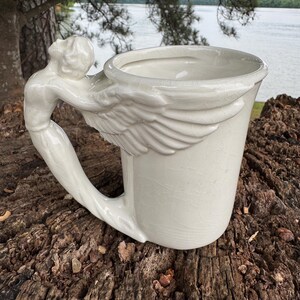 May include: A white ceramic mug with a sculpted angel handle and wing detail. The mug is set on a weathered wooden surface, with a lake and trees in the background. The mug has a capacity of approximately 12 ounces.