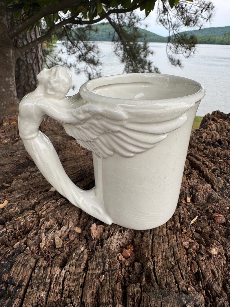 May include: A white ceramic mug with a sculpted angel handle and wing detail. The mug is set on a weathered wooden surface, with a lake and trees in the background. The mug has a capacity of approximately 12 ounces.