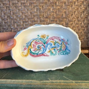 Könnte beinhalten: Eine ovale weiße Keramikschale mit gewelltem Rand, die in der Mitte mit einem dekorativen Motiv aus farbenfrohen, wirbelnden Akanthusblättern in Gelb, Rosa, Blau und Grün verziert ist, akzentuiert durch kleine florale Details in der Mitte.