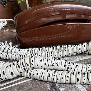 Puede incluir: Un teléfono marrón vintage con un cable blanco y negro moteado. El teléfono tiene un diseño clásico, con un auricular y una base curvados. El cable está enrollado y se encuentra delante del teléfono.