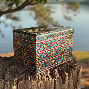 May include: A decorative, rectangular box with a vibrant floral design. The box features a red base with green and yellow floral patterns. The top and sides are adorned with intricate details, creating a colorful and ornate appearance. The box is placed on a weathered wooden surface.