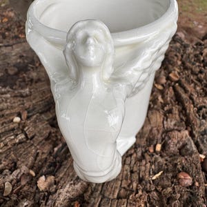 May include: A white ceramic vase with a sculpted angel figure on the side. The vase has a wide opening and a textured, crackled surface. The angel's face is detailed, and the wings are spread around the vase.