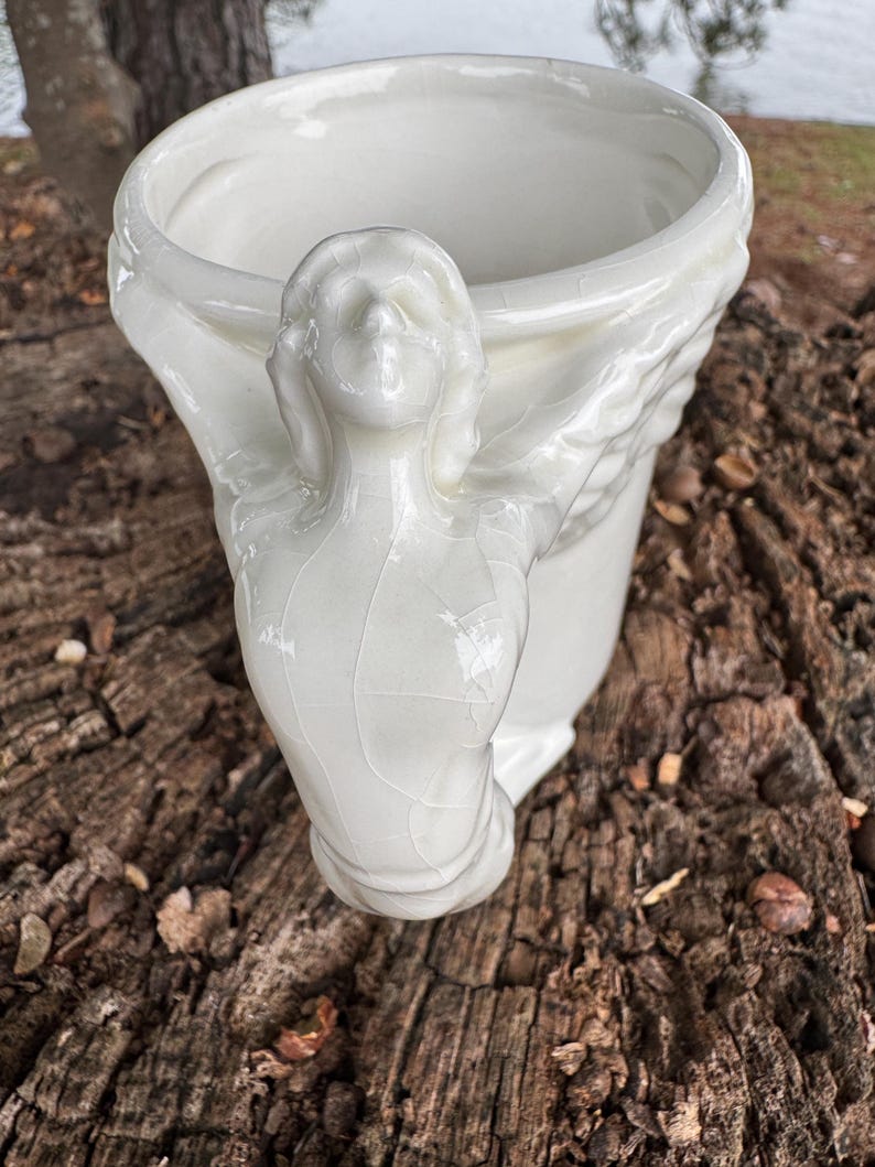 May include: A white ceramic vase with a sculpted angel figure on the side. The vase has a wide opening and a textured, crackled surface. The angel's face is detailed, and the wings are spread around the vase.