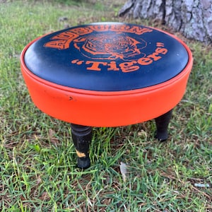 May include: A round, low stool with a navy blue padded top featuring the words "AUBURN" and "Tigers" in orange, along with a tiger graphic. The stool has an orange rim, black legs, and is set on grass.