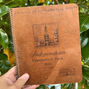 May include: A brown spiral-bound composition book with the word "Independence" and "Composition Book" printed on the cover. The cover features an illustration of a building and the Ennis logo. The book is held by a hand.