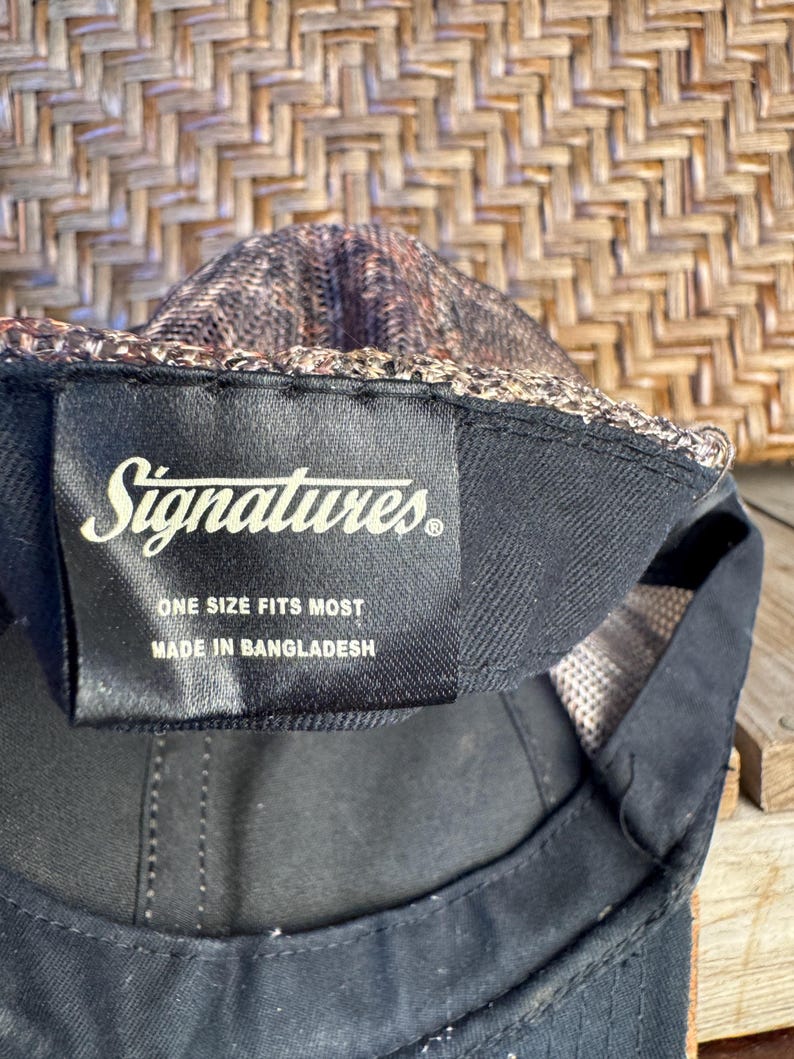 Puede incluir: Gorra de b&eacute;isbol negra con una parte superior de malla estampada y una visera negra. La etiqueta dice "Signatures" y "Talla &uacute;nica". Hecho en Bangladesh.