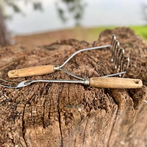 May include: Two vintage kitchen tools with wooden handles rest on a weathered tree stump. One is a potato masher, the other a fork-like utensil. The metal components are silver-colored, and the wood is light brown.