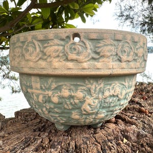 May include: A two-toned, round planter with a floral relief design. The upper section is a light tan color, while the lower section is a muted green. The planter has a small drainage hole and sits on three small feet. Ideal for outdoor or indoor use.