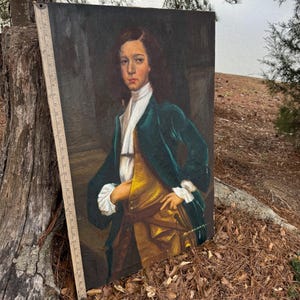 Pode incluir: Uma pintura de retrato antigo de uma pessoa jovem com uma jaqueta de veludo verde, colete dourado e camisa branca com babados. A pintura &eacute; definida contra um fundo escuro, com uma r&eacute;gua vis&iacute;vel ao longo da borda. O sujeito tem cabelo castanho comprido e est&aacute; olhando diretamente para o espectador.