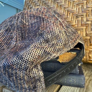 Puede incluir: Gorra de b&eacute;isbol de malla con una correa ajustable negra. La gorra presenta un patr&oacute;n de camuflaje en tonos marrones, negros y beige. El sombrero est&aacute; sobre una pila de libros antiguos y una superficie de madera.