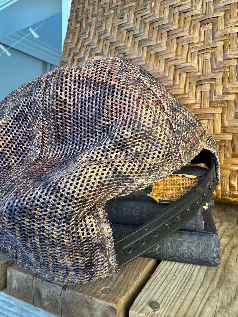 Puede incluir: Gorra de b&eacute;isbol de malla con una correa ajustable negra. La gorra presenta un patr&oacute;n de camuflaje en tonos marrones, negros y beige. El sombrero est&aacute; sobre una pila de libros antiguos y una superficie de madera.