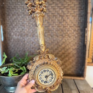 Puede incluir: Reloj de pared dorado ornamentado con diseño floral y esfera redonda. El reloj tiene un tallo largo y curvo y es sostenido por una mano. La esfera presenta números romanos negros y manecillas. El reloj está hecho de un material dorado.