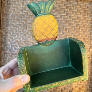May include: A dark green wooden napkin holder featuring a carved pineapple accent on the back panel. The pineapple is painted in shades of yellow and orange with green leaves at the top. The base and sides have a distressed finish with visible yellow.