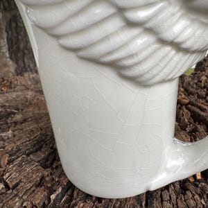 May include: Cream-colored ceramic mug with a wing-shaped handle. The mug features a textured, cracked glaze and a sculpted wing detail on the side. The mug is designed for beverages and has a classic, elegant aesthetic.