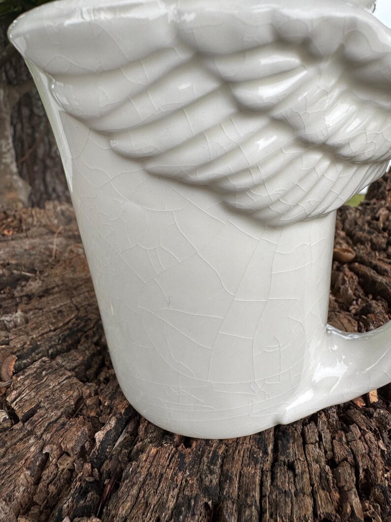 May include: Cream-colored ceramic mug with a wing-shaped handle. The mug features a textured, cracked glaze and a sculpted wing detail on the side. The mug is designed for beverages and has a classic, elegant aesthetic.