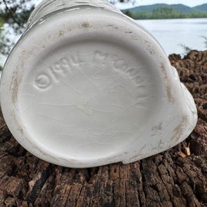 May include: A white ceramic pitcher with a textured surface and a cracked glaze. The bottom of the pitcher features embossed text and a circular logo. The pitcher is sitting on a weathered wooden surface.