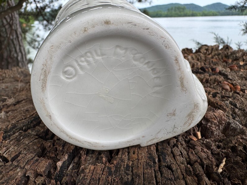 May include: A white ceramic pitcher with a textured surface and a cracked glaze. The bottom of the pitcher features embossed text and a circular logo. The pitcher is sitting on a weathered wooden surface.