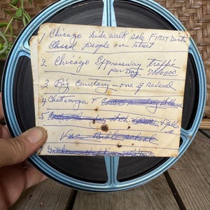 Puede incluir: Una bobina de película vintage con un borde azul y una nota manuscrita adjunta. La nota enumera varios temas, incluyendo "Chicago side walk sale" y "Chicago Expressway Traffic". La bobina se sostiene contra una superficie de madera.