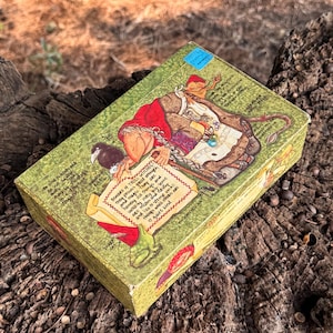 May include: A rectangular green box featuring a detailed illustration of a troll witch with a red hood, a black bird, and a frog. The box includes handwritten-style text about troll witches, a small blue sticker, and the text Enchanted Art.