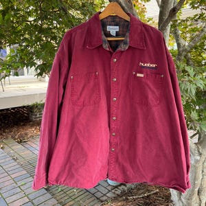May include: A maroon Carhartt long-sleeve button-down shirt featuring a plaid flannel lining, a pointed collar, and two chest pockets. The left chest pocket displays the embroidered text "hueber" in yellow and blue lettering above the pocket flap.