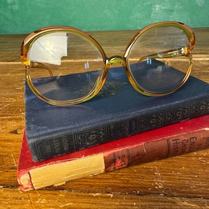 May include: Oversized eyeglasses with a translucent amber-colored frame and clear lenses. The frame features a rounded shape with a distinct bridge. The inner temple arm is clearly marked with the text "MADE IN GERMANY" and additional small characters.