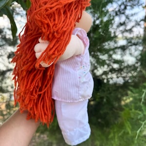 Puede incluir: Una muñeca hecha a mano con cabello de hilo naranja brillante. La muñeca lleva una camiseta a rayas rosas y blancas y pantalones blancos. Los brazos de la muñeca están extendidos y está de espaldas a la cámara.