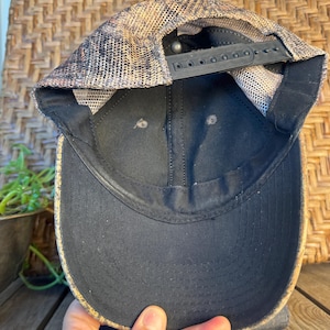 Puede incluir: Gorra de b&eacute;isbol negra con visera curva. La gorra tiene una parte trasera de malla en tonos marrones y beige, y una correa negra ajustable. La parte inferior de la visera es negra y la gorra est&aacute; sujeta por una mano.