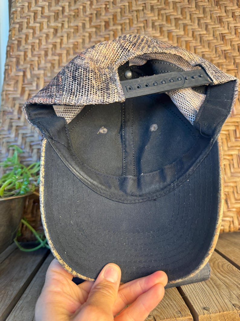 Puede incluir: Gorra de b&eacute;isbol negra con visera curva. La gorra tiene una parte trasera de malla en tonos marrones y beige, y una correa negra ajustable. La parte inferior de la visera es negra y la gorra est&aacute; sujeta por una mano.