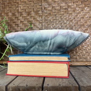 Op de afbeelding: Een grote, ovale keramische kom met een blauw en grijs glazuur staat op twee boeken. De boeken hebben rode en blauwe covers. De kom is een decoratief stuk.