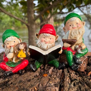 Op de afbeelding: Drie keramische tuinkabouters met rode, groene en witte kleding. Eén kabouter houdt een kleine pop vast, een ander leest een boek en de derde houdt een kleine boot vast. Ze zitten op een stuk hout.