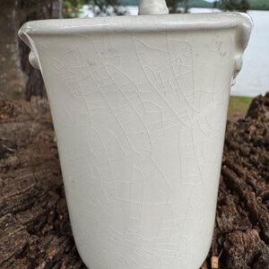 May include: A white ceramic container with a lid, featuring a crackle glaze finish. The container has a cylindrical shape with a slightly flared rim and small handles. The lid has a small knob.
