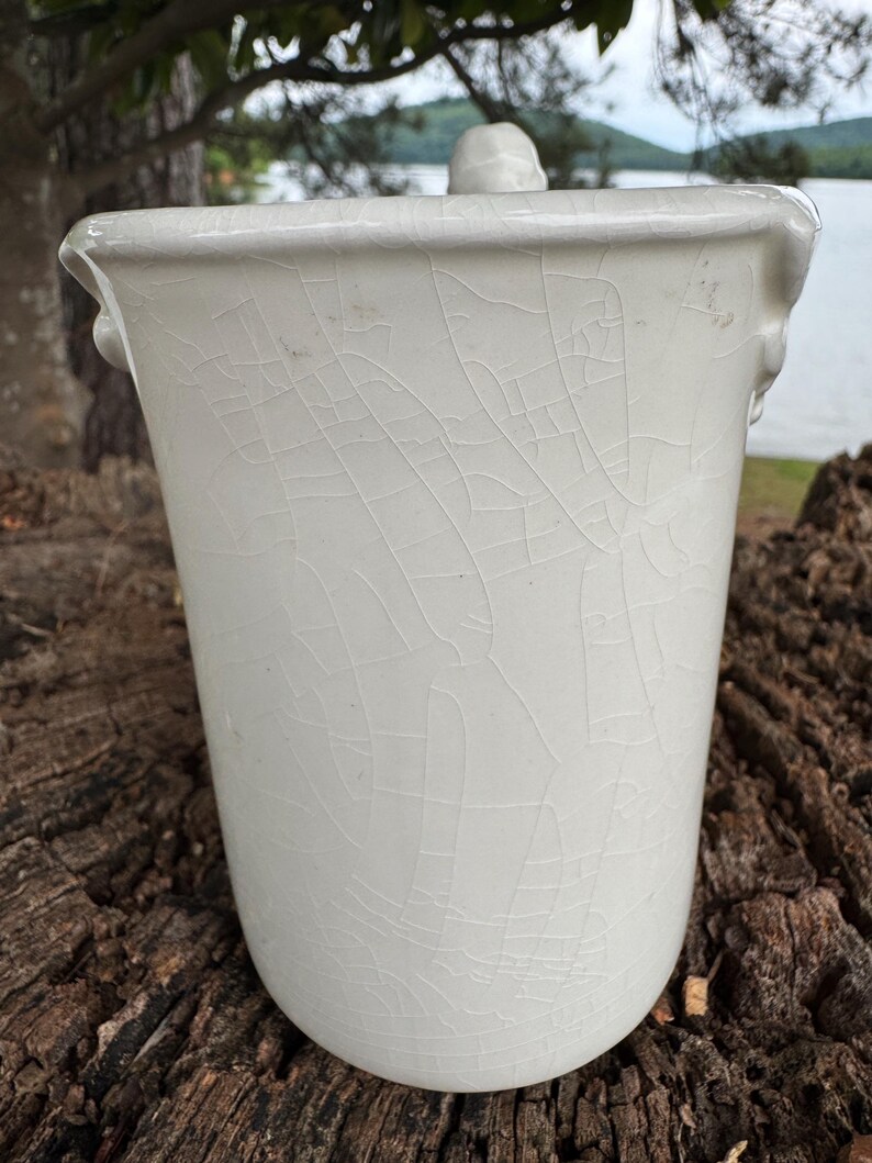 May include: A white ceramic container with a lid, featuring a crackle glaze finish. The container has a cylindrical shape with a slightly flared rim and small handles. The lid has a small knob.