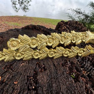 Può includere: Due braccialetti in metallo color oro con disegni floreali e di foglie. I braccialetti sono appoggiati su una superficie di legno marrone scuro e strutturato. I braccialetti hanno un aspetto dettagliato e ornato.