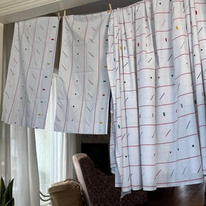 Puede incluir: Cortinas blancas con un patrón repetitivo de líneas horizontales rojas, líneas diagonales negras y pequeños cuadrados de colores. Las cortinas cuelgan de una barra de madera con pinzas. El estilo es retro y minimalista.