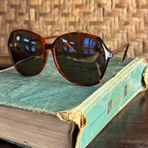 May include: Oversized sunglasses with a glossy brown tortoiseshell frame and dark tinted lenses. The glasses feature a rounded, slightly angular shape with thin temples and are positioned on top of a weathered, teal-coloured hardcover book.