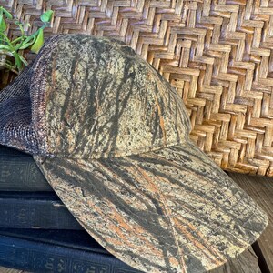Puede incluir: Gorra de b&eacute;isbol de camuflaje con visera curva. La gorra presenta una parte trasera de malla y un patr&oacute;n marr&oacute;n, beige y negro que se asemeja a un bosque. El sombrero est&aacute; sobre una pila de libros azul oscuro.