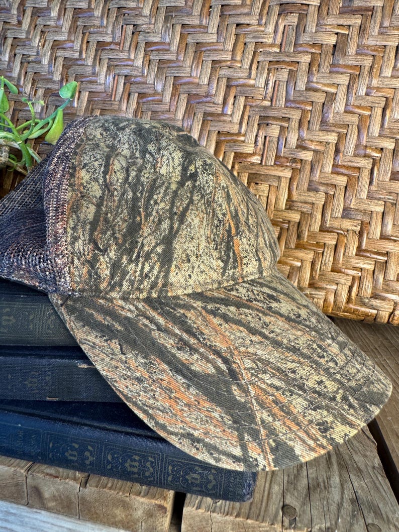 Puede incluir: Gorra de b&eacute;isbol de camuflaje con visera curva. La gorra presenta una parte trasera de malla y un patr&oacute;n marr&oacute;n, beige y negro que se asemeja a un bosque. El sombrero est&aacute; sobre una pila de libros azul oscuro.