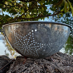 May include: A vintage, silver-toned metal colander with numerous small holes. The colander has two handles and three small feet. The image is taken outdoors, with a blurred background of trees and water.