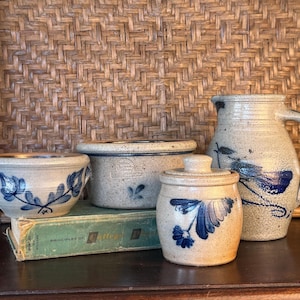May include: A collection of four stoneware pieces featuring a speckled beige finish and hand-painted cobalt blue floral motifs. The set includes a small handled bowl, a wide basin, a lidded jar, and a pitcher with a curved handle and spout.