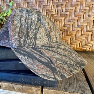 Puede incluir: Una gorra de b&eacute;isbol de camuflaje con la parte trasera de malla. La gorra presenta un patr&oacute;n marr&oacute;n, verde y negro. El sombrero est&aacute; apoyado en una pila de tres libros azul oscuro.