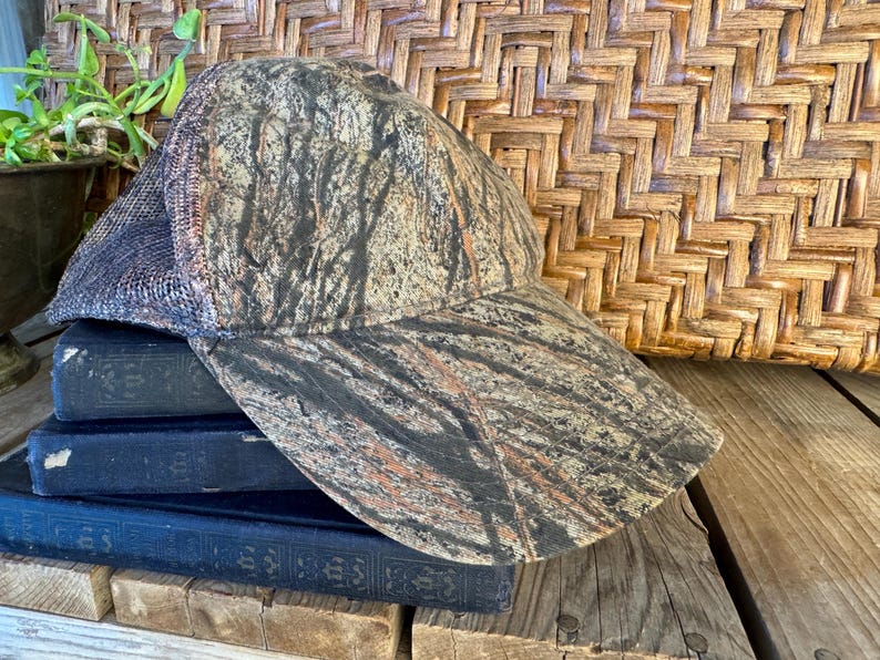 Puede incluir: Una gorra de b&eacute;isbol de camuflaje con la parte trasera de malla. La gorra presenta un patr&oacute;n marr&oacute;n, verde y negro. El sombrero est&aacute; apoyado en una pila de tres libros azul oscuro.