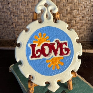 May include: A circular ceramic wall plaque with a scalloped edge and a top hanging loop. The center features a light blue background with the word LOVE in raised red letters, flanked by two yellow sun icons. The plaque has a glossy cream finish.