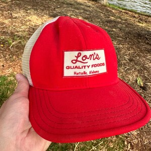 Puede incluir: Gorra de camionero roja y blanca de malla con un parche blanco que dice "Lon's Quality Foods Hartselle, Alabama".