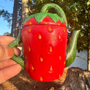 May include: A red ceramic teapot shaped like a strawberry with green leaves and yellow seeds. The teapot has a handle and a spout.