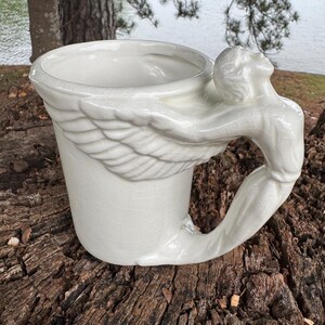 May include: White ceramic mug with a unique design. The handle is formed by a sculpted figure with outstretched arms and wings. The mug has a crackled glaze finish, adding a vintage aesthetic. The mug is sitting on a wooden surface.