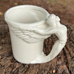 May include: Cream-colored ceramic mug with a sculpted handle featuring a figure with outstretched arms and wings. The mug has a cylindrical shape and a slightly flared rim. The handle is integrated into the wing design.
