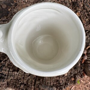 May include: A white ceramic planter with a rounded bowl shape and a small handle. The planter has a crackled glaze finish and is set against a natural wood background. The planter is empty.